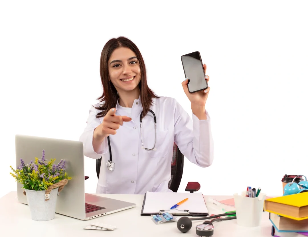 Médica sorridente sentada à mesa com laptop e estetoscópio, segurando um celular na mão direita e apontando para frente com a esquerda.