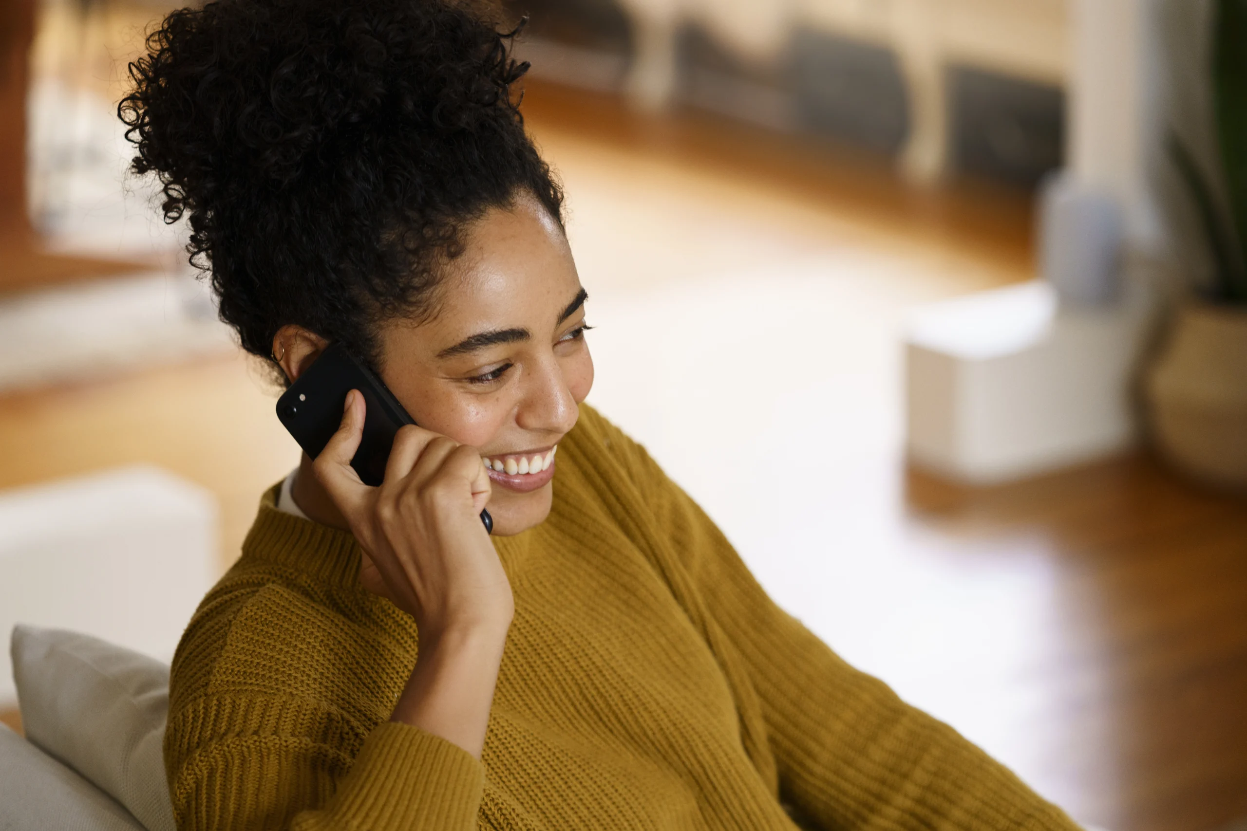 Mulher sorridente falando ao telefone celular enquanto está sentada em casa, em um ambiente acolhedor e iluminado.