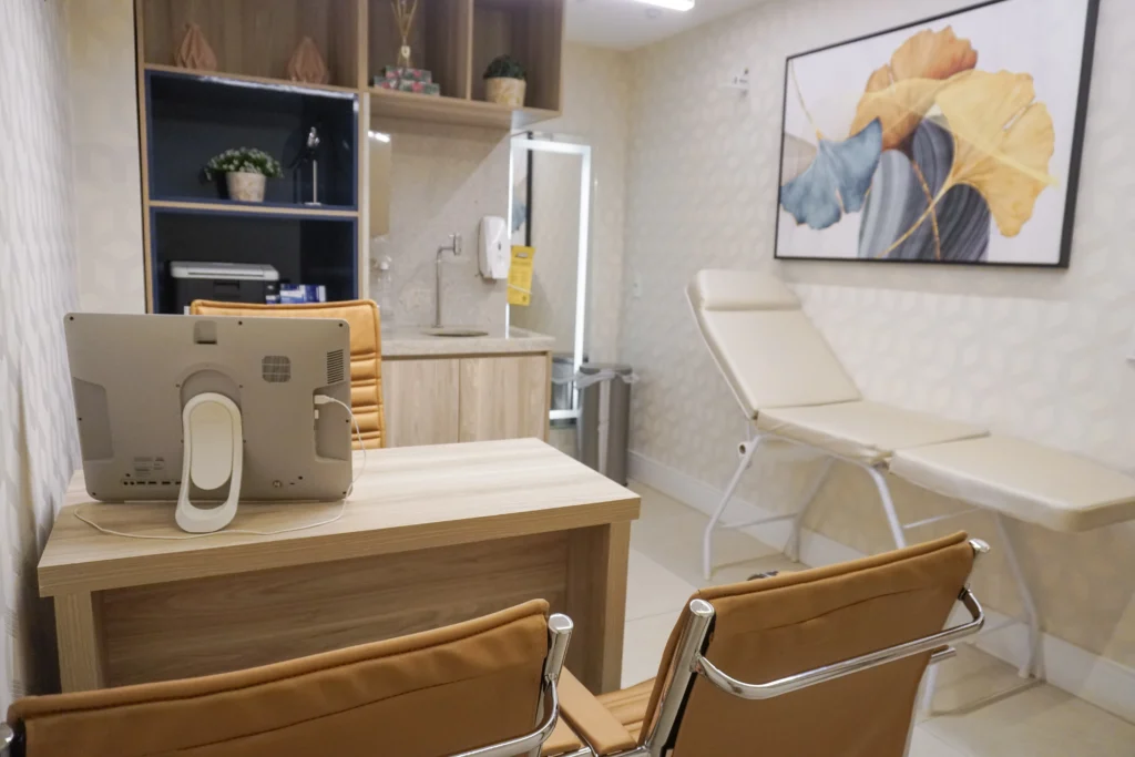 Interior de consultório médico visto de frente, com mesa de trabalho, cadeiras confortáveis, estante planejada, pia de apoio e maca posicionada ao lado de um quadro decorativo.