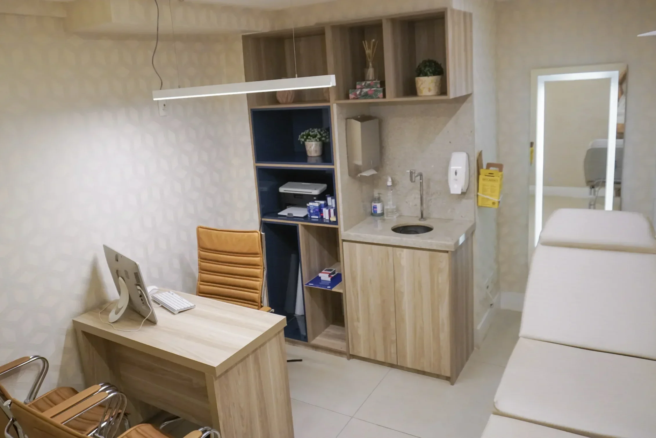 Consultório médico com mesa de atendimento em madeira, cadeiras em couro caramelo, armários planejados, pia embutida e maca ao fundo, criando um ambiente organizado e confortável.