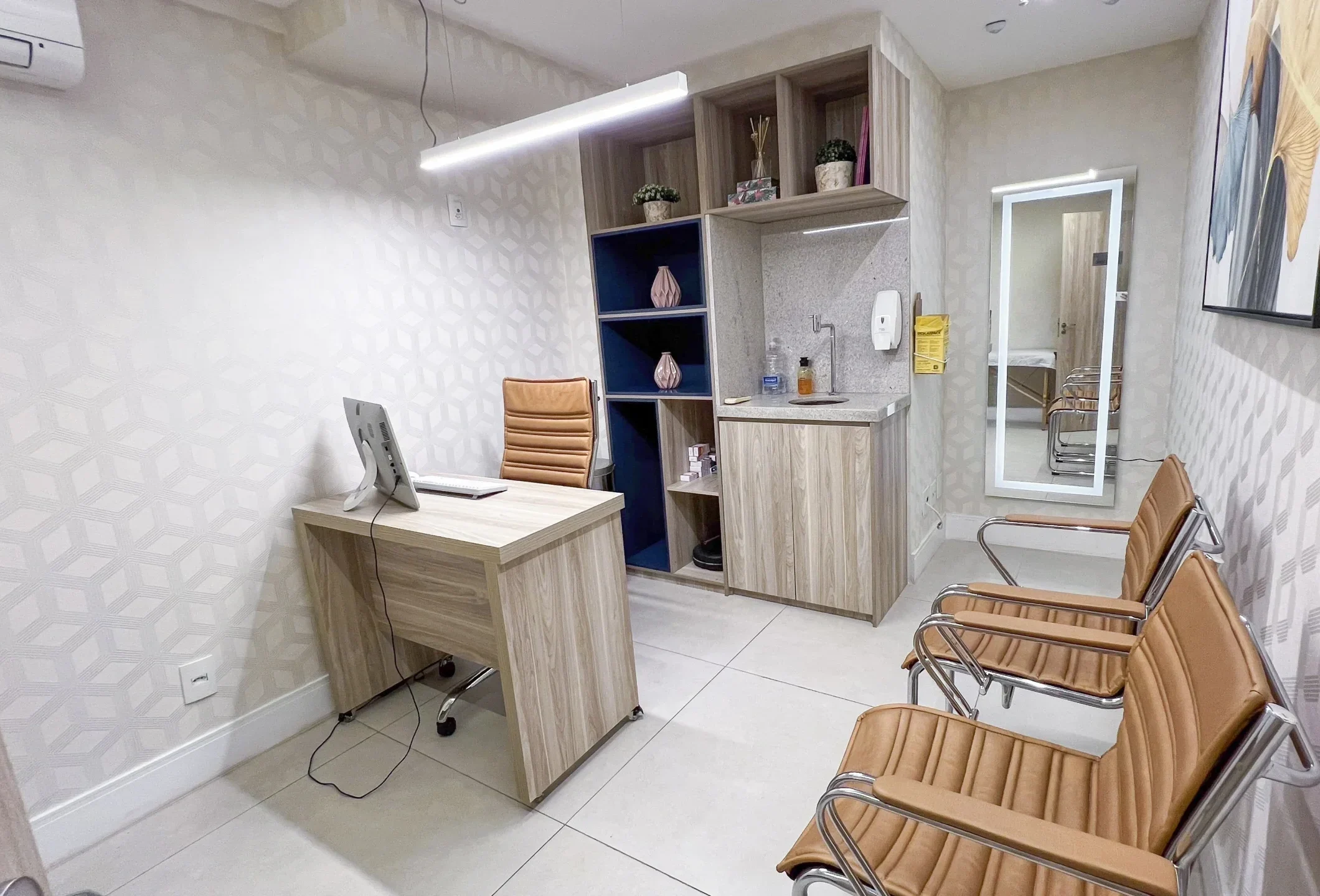 Consultório médico moderno com mesa de atendimento em madeira, cadeira executiva caramelo, cadeiras de espera combinando, estante planejada com objetos decorativos, pia embutida e espelho ao fundo, em ambiente claro e organizado.