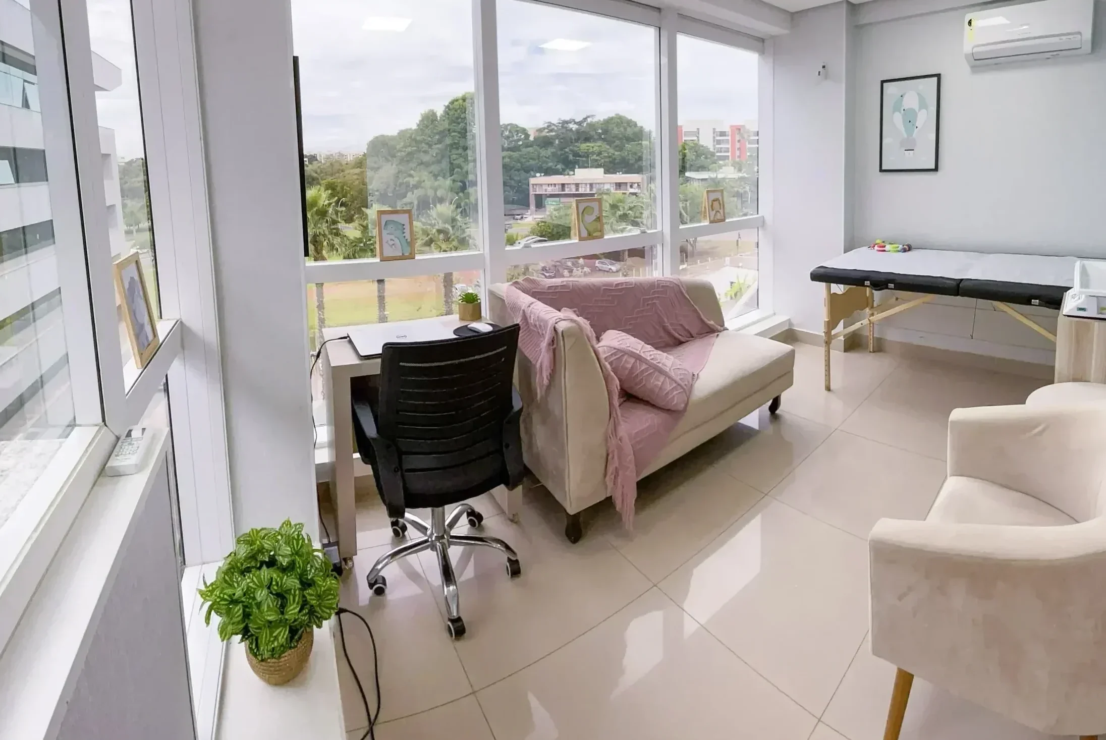 Consultório médico moderno com mesa de atendimento, cadeiras caramelo, estante com objetos decorativos, pia integrada e maca de exames ao fundo, em ambiente bem iluminado.