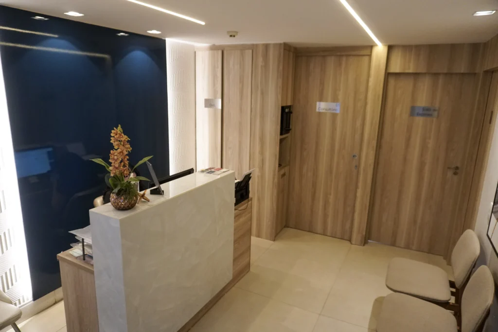 Recepção de clínica médica com balcão claro, painel azul elegante, iluminação embutida e portas de consultório em madeira clara, transmitindo ambiente moderno e acolhedor.